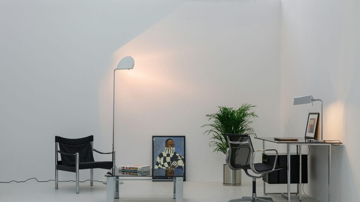 Minimalist white studio interior with chrome coffee table, black canvas chair, floor lamp, Eames-style task chair, and palm plant
